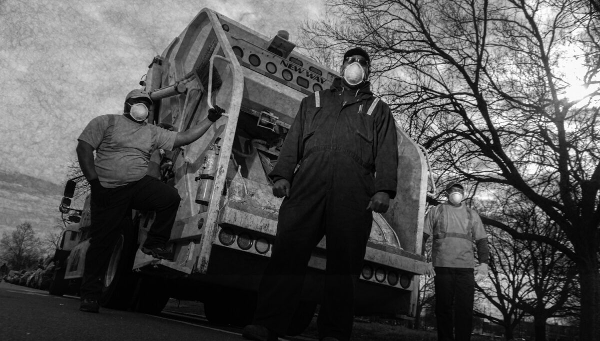 3 sanitation workers stand in front of a garbage truck