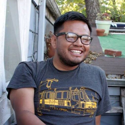 Rigo sitting on steps and leaning back on his left arm. He is wearing black glasses and a grey T-shirt. He is smiling.