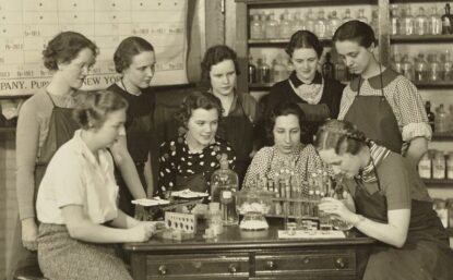 group of women standing around a lab table with periodic table in background