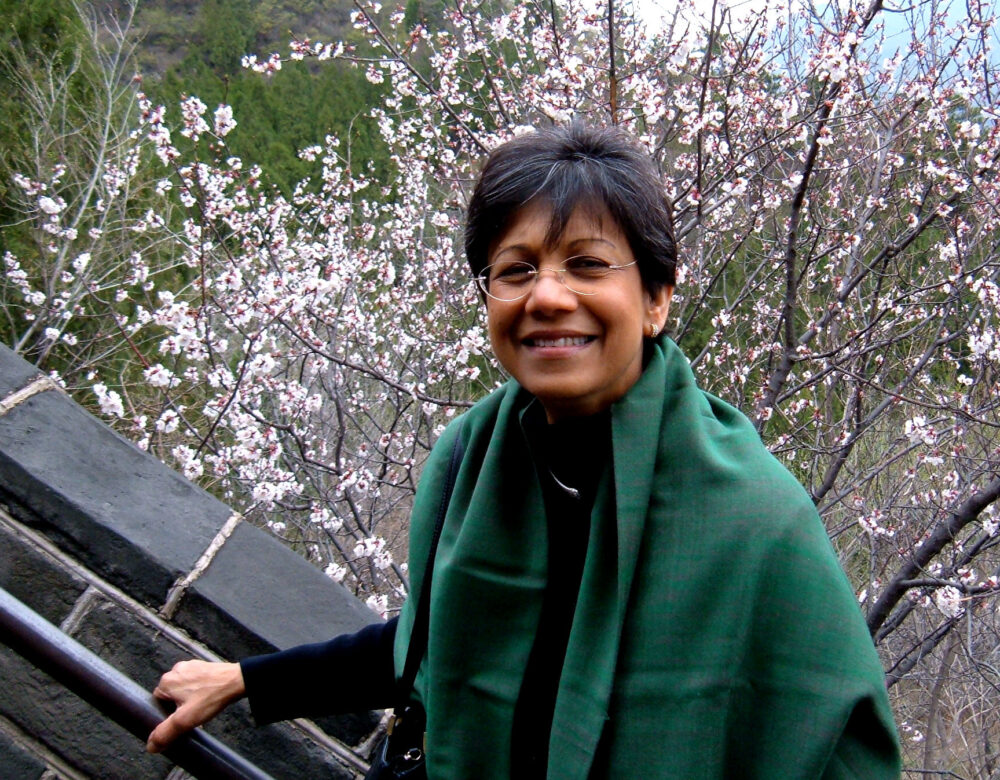 Uma Chowdhry wearing green shawl and glasses, hand on a rail, cherry blossoms in background