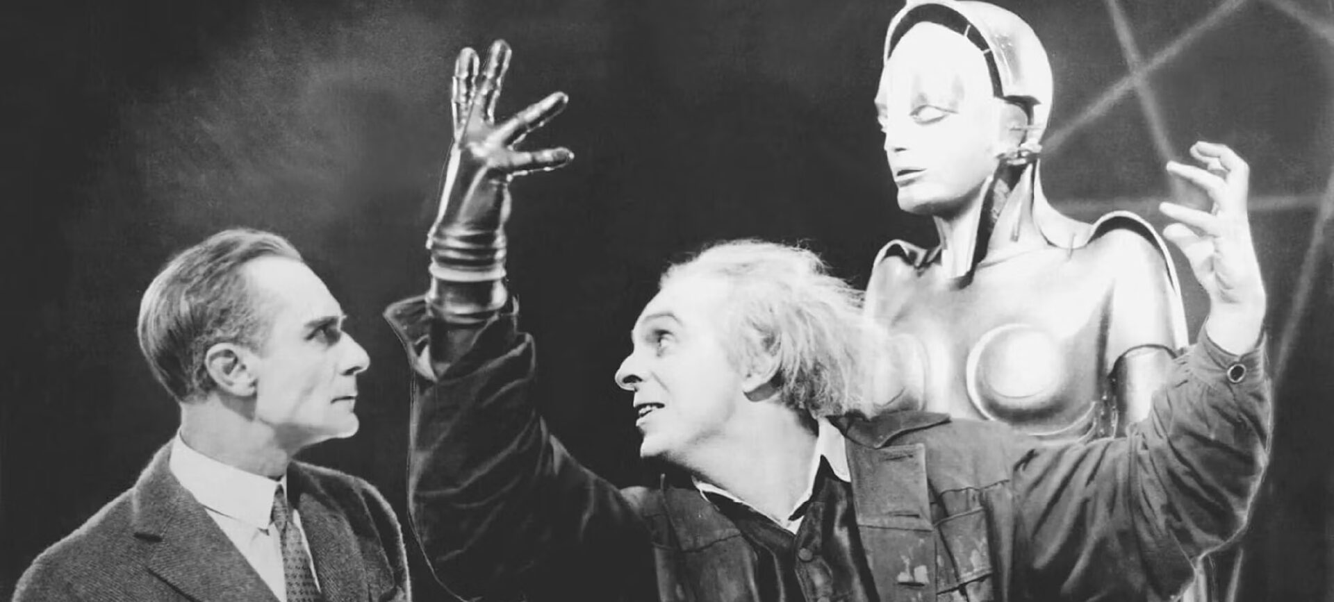 black and white film still with two men, one with white hair and hand raised, in front of a mannequin